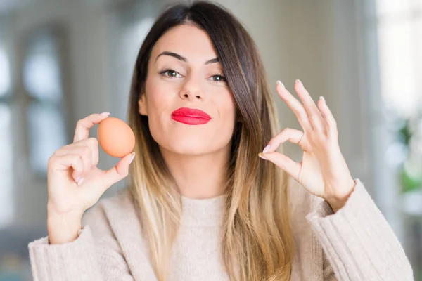 年轻美丽的女人拿着新鲜的鸡蛋在家里做确定的标志与手指 优秀的象征 — 图库照片
