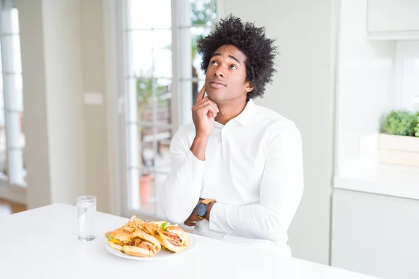 African American hungry man eating hamburger for lunch with hand on chin thinking about question, pensive expression. Smiling with thoughtful face. Doubt concept.