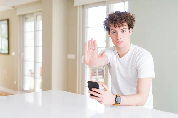 Young man using smartphone at home with open hand doing stop sign with serious and confident expression, defense gesture
