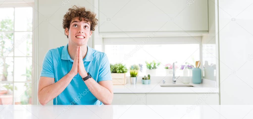 Wide shot of young handsome man at home praying with hands together asking for forgiveness smiling confident.