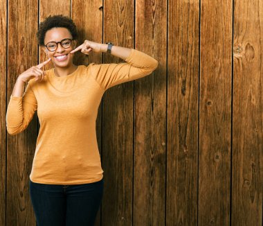 Kendinden emin gösterilen ve işaret parmakları diş ve ağız ile gülümseyen izole arka plan üzerinde gözlük takan genç güzel Afrikalı-Amerikalı kadın. Sağlık kavramı.