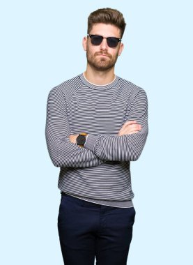 Çapraz kolları olan yüzündeki güneş gözlüğü şüpheci ve sinir, onaylamayan ifade giyen genç yakışıklı adam. Negatif kişi.
