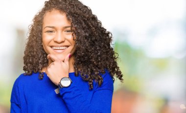 Genç güzel kadın kıvırcık saçlı emin çapraz kollar ve çeneme kaldırdı el ile gülümseme ile kameraya bakarak kış kazak giyiyor. Pozitif düşünce.