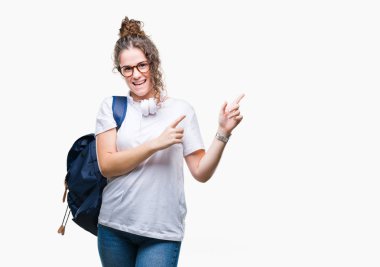 Gülümseyen ve iki elleri ve parmakları ile tarafına işaret kamera bakarak izole arka plan üzerinde sırt çantası ve kulaklıklar giyen genç esmer öğrenci kız.