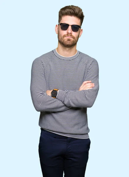 Çapraz kolları olan yüzündeki güneş gözlüğü şüpheci ve sinir, onaylamayan ifade giyen genç yakışıklı adam. Negatif kişi.