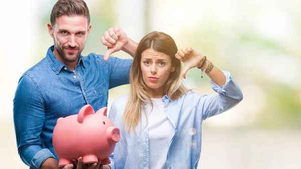Young couple in love holding piggy bank over isolated background with angry face, negative sign showing dislike with thumbs down, rejection concept