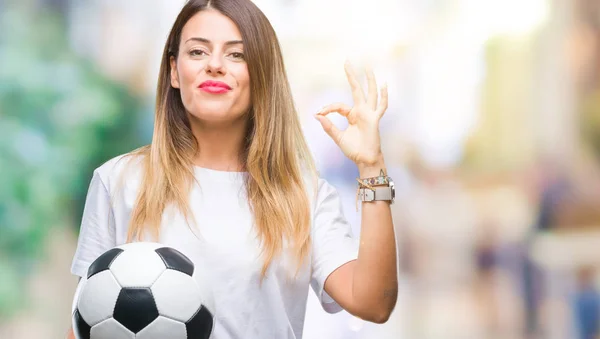 Young beautiful woman holding soccer ball over isolated background doing ok sign with fingers, excellent symbol