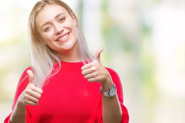 Young blonde woman wearing winter sweater over isolated background approving doing positive gesture with hand, thumbs up smiling and happy for success. Looking at the camera, winner gesture.