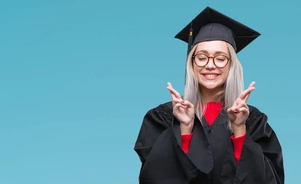 若いブロンドの女性は 希望と目を閉じて交差指の笑みを浮かべて隔離された背景に大学院の制服を着ています 運と迷信的な概念 — ストック写真