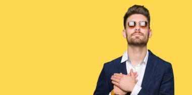 Güneş gözlüğü ve ile gülümseyen moda ceket giyen genç yakışıklı zarif adam kapalı gözler ve yüzünde minnettar jest ile göğüs üzerinde eller. Sağlık kavramı.