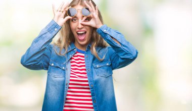 Güzel genç sarışın kadın gülümsüyor, parmakları ile mutlu yüz üzerinden seyir göz el ile ok jest yapıyor izole arka plan üzerinde güneş gözlüğü takıyor.