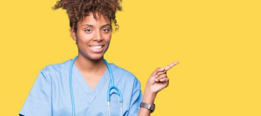 Kameraya bakarak yan el ve parmak ile işaret eden genç Afro-Amerikan doktor kadın yüzünde kocaman bir gülümseme ile izole arka plan üzerinde.