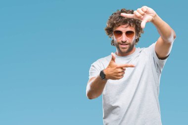 Yakışıklı İspanyol modeli adam elleriyle yapım çerçeve ve parmakları ile mutlu yüz gülümseyen izole arka plan üzerinde güneş gözlüğü takıyor. Yaratıcılık ve Fotoğrafçılık kavramı.