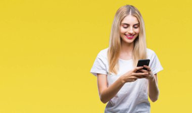 Ayakta ve diş gösterilen kendine güvenen bir gülümsemeyle gülümseyerek mutlu bir yüz ile izole arka plan üzerinde akıllı telefon kullanan genç güzel sarışın kadın