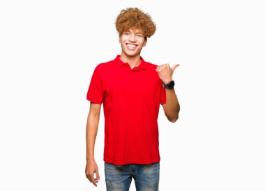 Kırmızı giyen afro saçlı genç yakışıklı adam mutlu ile gülümseyen t-shirt arıyorsunuz ve başparmak yanında işaret eden oktan yukarı yüz.