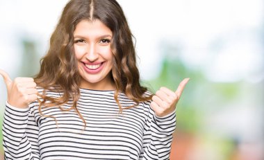 Çizgili süveter başarı işareti el ile pozitif jest yapıyor giyen genç güzel kadın başparmak gülümseyen ve mutlu. Neşeli bir ifadeyle, kazanan jest kameraya bakıyor.