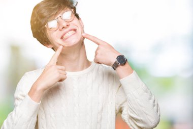 Kendinden emin gösterilen ve işaret parmakları diş ve ağız ile gülümseyen izole arka plan üzerinde gözlük takan yakışıklı delikanlı. Sağlık kavramı.