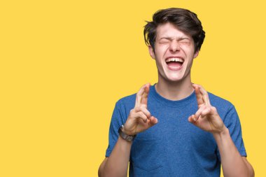 Umut ve gözleri kapalı geçiş parmaklarıyla gülümseyen izole arka plan üzerinde mavi t-shirt giyen genç yakışıklı adam. Şans ve batıl kavramı.