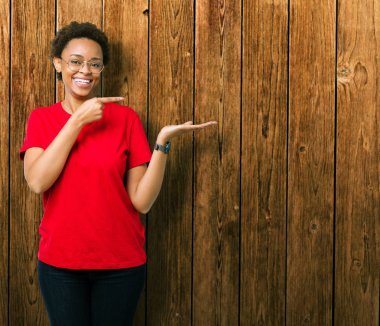 Güzel genç Afro-Amerikan kadın hayrete ve el ile sunulması ve parmak ile işaret ederken kameraya gülümseyen izole arka plan üzerinde gözlük.