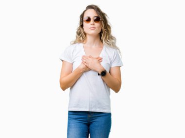 Güzel genç sarışın kadın ile gülümseyen izole arka plan üzerinde güneş gözlüğü takıyor kapalı gözler ve yüzünde minnettar jest ile göğüs üzerinde eller. Sağlık kavramı.