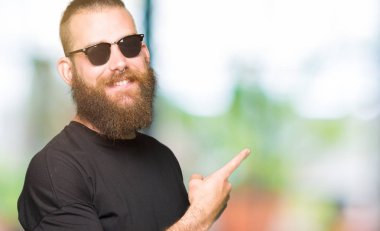 Yüzünde, kocaman bir gülümseme ile güneş gözlüğü takmış genç hipster adam kameraya bakarak yan el ve parmak ile işaret.