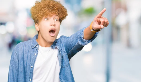 Young handsome man with afro hair wearing denim jacket Pointing with finger surprised ahead, open mouth amazed expression, something in front