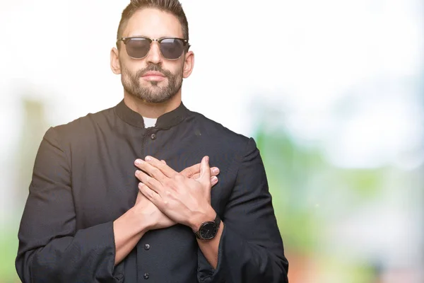 Young Christian priest wearing sunglasses over isolated background smiling with hands on chest with closed eyes and grateful gesture on face. Health concept.