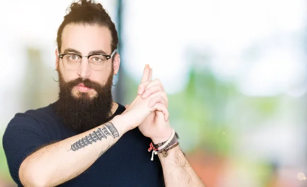 Young hipster man with long hair and beard wearing glasses Holding symbolic gun with hand gesture, playing killing shooting weapons, angry face