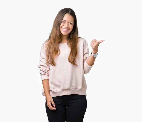 年轻美丽的黑发女人穿着粉红色的运动衫在孤立的背景微笑着与快乐的脸看 用拇指指着一边 — 图库照片