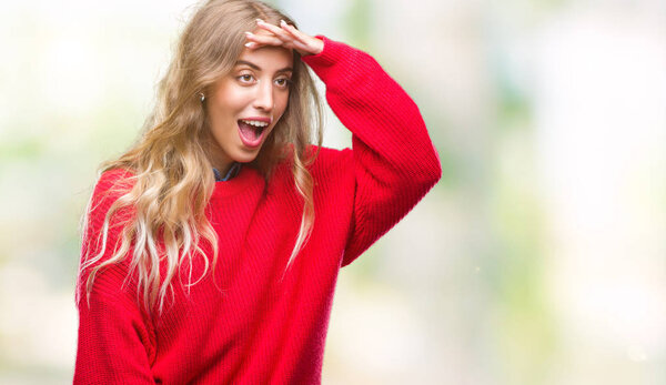 Beautiful young blonde woman wearing winter sweater over isolated background very happy and smiling looking far away with hand over head. Searching concept.