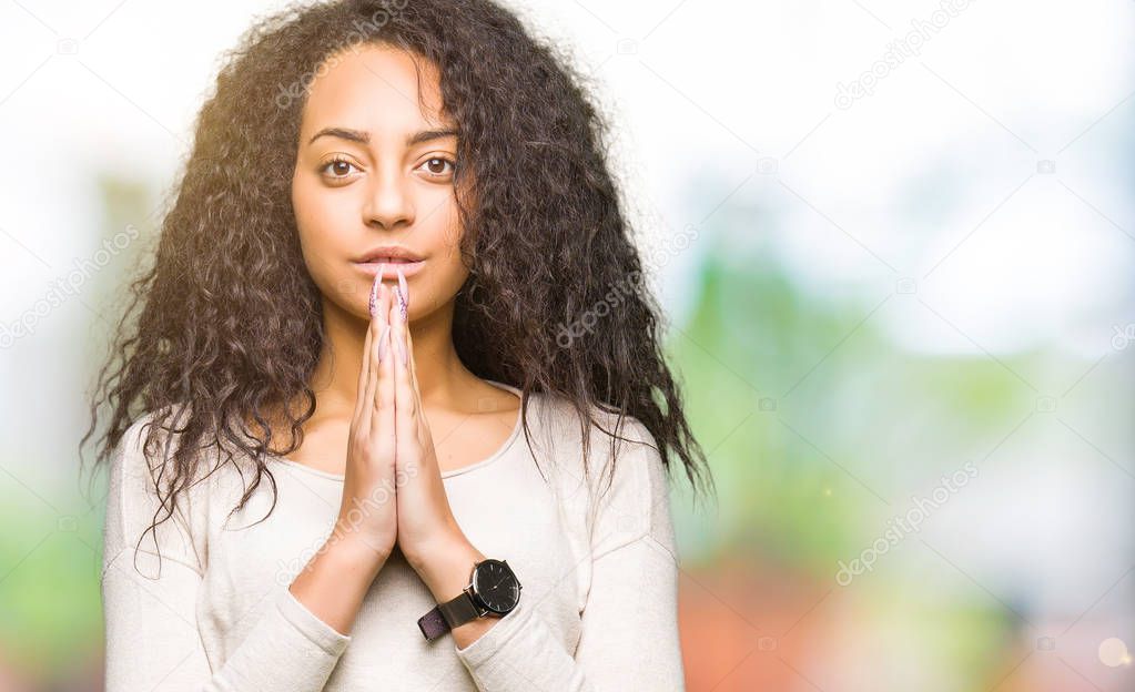 Young beautiful girl with curly hair wearing casual sweater praying with hands together asking for forgiveness smiling confident.