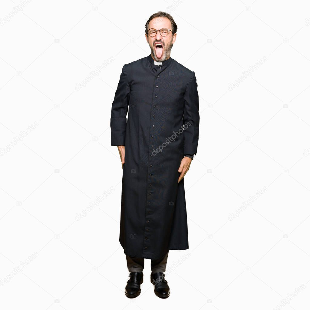 Middle age priest man wearing catholic robe sticking tongue out happy with funny expression. Emotion concept.