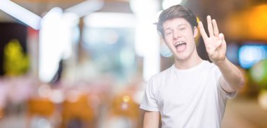 Gösterilen ve parmakları ile işaret izole arka plan üzerinde rahat beyaz t-shirt giyen genç yakışıklı adam kendinden emin ve mutlu gülümseyerek iken üçüncü.