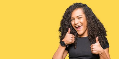 Genç güzel kadın kıvırcık saçlı başarı işareti el ile pozitif jest yapıyor başparmak gülümseyen ve mutlu. Neşeli bir ifadeyle, kazanan jest kameraya bakıyor.