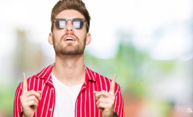 Yakışıklı delikanlı güneş gözlüğü takıyor hayrete ve yukarıya bakıyor ve parmakları ile işaret ve silah kaldırdı şaşırttı.