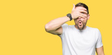 Yakışıklı adam utanç ifadesi ile parmaklarımın arasından seyir yüz ve gözler el ile kapsayan şok bakmak rahat beyaz t-shirt giyen.