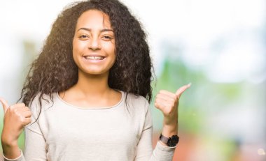 El ile olumlu hareketi yaparken rahat kazak başarı işareti giyiyor kıvırcık saçlı genç güzel kız gülümseyerek ve mutlu Yaşasın. Neşeli bir ifadeyle, kazanan jest kameraya bakıyor.