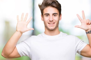 Gösterilen ve parmakları ile işaret izole arka plan üzerinde beyaz t-shirt giyen genç yakışıklı adam kendinden emin ve mutlu gülümseyerek süre sekiz numara.