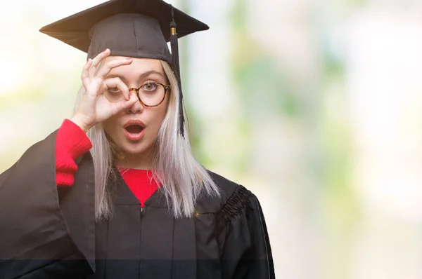 年轻的金发女子穿毕业制服在孤立的背景做 手势震惊与惊讶的脸 眼睛看通过手指 不信表达 — 图库照片