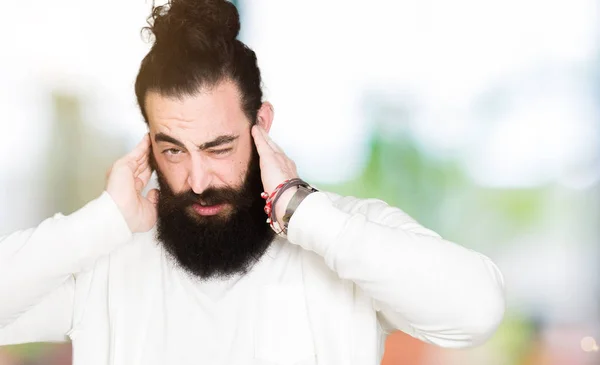 Young man with long hair and beard wearing sporty sweatshirt covering ears with fingers with annoyed expression for the noise of loud music. Deaf concept.