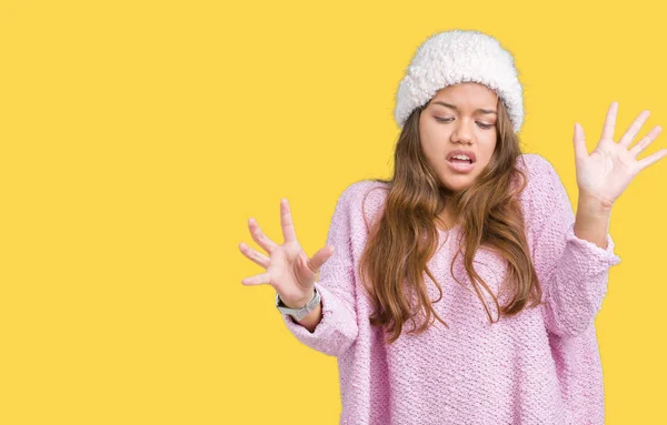 Young beautiful brunette woman wearing sweater and winter hat over isolated background afraid and terrified with fear expression stop gesture with hands, shouting in shock. Panic concept.