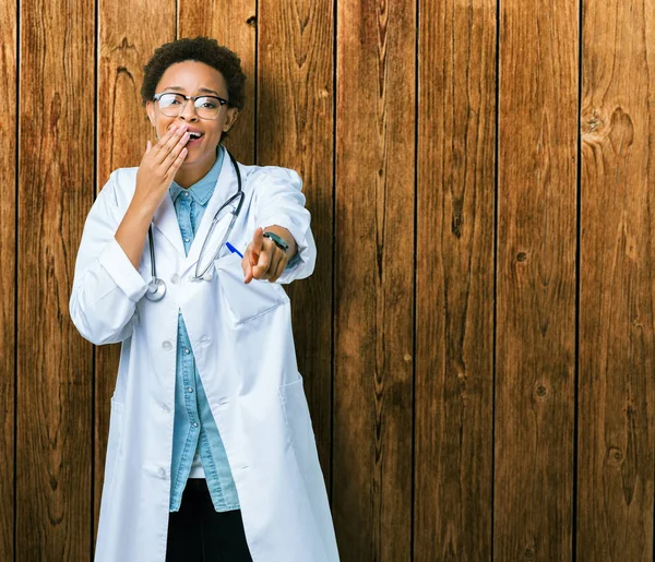 Young african american doctor woman wearing medical coat over isolated background Laughing of you, pointing to the camera with finger hand over mouth, shame expression