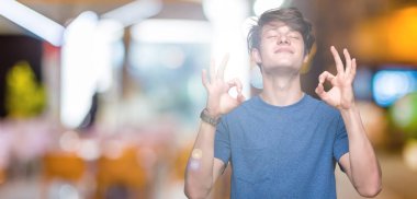 İzole arka plan üzerinde mavi t-shirt giyen genç yakışıklı adam rahatla ve gülümseyen gözlerle kapalı parmak jestle meditasyon yapıyor. Yoga kavramı.