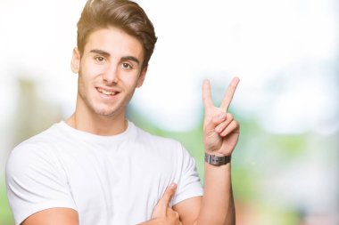 Mutlu yüz zafer işareti yaparken kameraya göz kırpıyor ile gülümseyen izole arka plan üzerinde beyaz t-shirt giyen genç yakışıklı adam. İki numaralı.