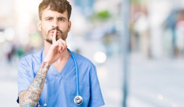 Genç yakışıklı dudaklarında parmak ile sessiz olmanı izole arka plan üzerinde cerrah üniforma giyen adam hemşire. Sessizlik ve gizli kavramı.