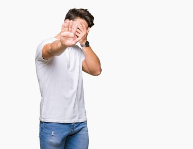 Giyen genç yakışıklı adam elleriyle gözlerini kapsayan izole arka plan üzerinde beyaz t-shirt ve stop jest ile acı ve korku ifadesi. Utandım ve negatif kavramı.