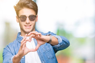 Genç yakışıklı adam aşık kalp simgesi ve elleriyle şekil gösteren gülümseyen izole arka plan üzerinde güneş gözlüğü takıyor. Romantik kavramı.