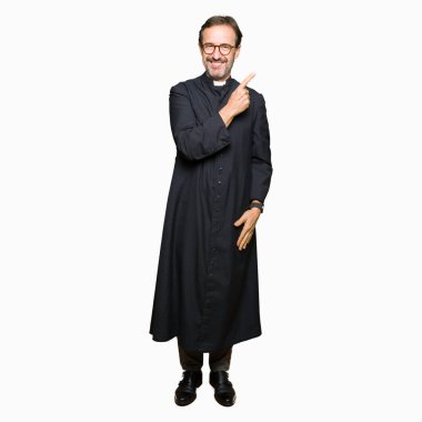 Orta yaş rahip adam Katolik bornoz neşeli bir gülümseme ile el ve parmak kadar yan yüzünde mutlu ve doğal ifade ile işaret yüz giymiş