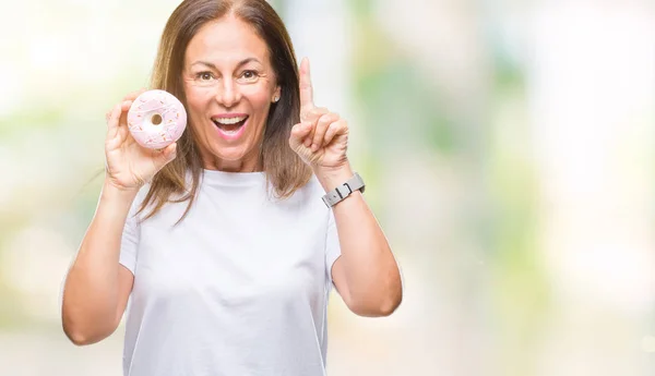 中年のヒスパニック系女性でピンクのドーナツを食べて分離アイデアに驚く背景や幸せそうな顔 ナンバーワンと人差し指の質問 — ストック写真