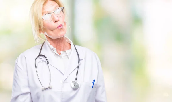 Middle age blonde doctor woman over isolated background making fish face with lips, crazy and comical gesture. Funny expression.
